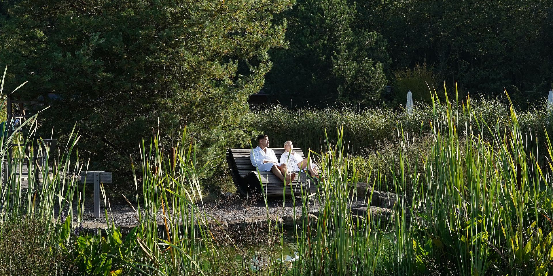 EIn Mann und eine Frau sitzen auf einem Liegestuhl aus Holz und schauen auf den Naturbadeteich